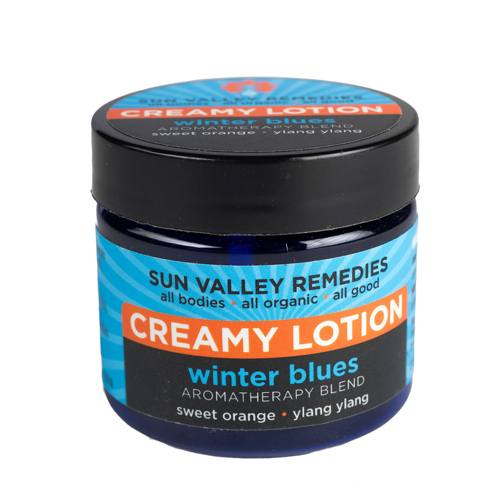 Two ounce cobalt jar of Winter Blues  creamy lotion. The blue label indicates the aromatherapy is sweet orange and ylang ylang